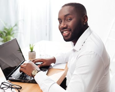 African American businessman happily looking in camera working on his laptop in office