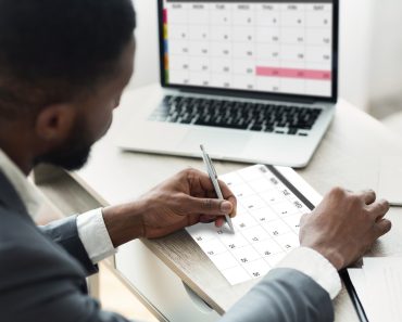 Black Businessman Using Calendar Program On Laptop And Writing Notes To Planner